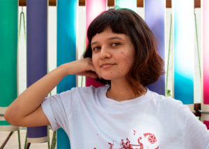 Na foto Rayanna está de cabelo curto, com pontas coloridas, ela veste uma blusa branca com detalhes vermelhos, atrás dela tem um fundo colorido, é um instrumento musical feito com canos nas cores verde, roxo, azul e rosa. Ela está sorrindo e faz um pose encostando a mão no rosto.