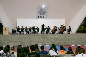 Apresentação da camerata de cordas da UFC. Foto colrida mostra o grupo no palco da concha acústica da UFC. Todos vestem perto. em primeiro plano aparece parte da plateia.